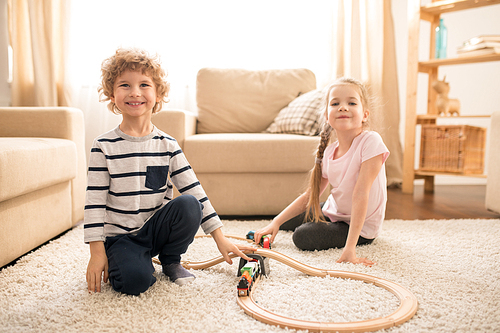 Two happy cute kids in casualwear sitting on soft carpet in the middle of living room while playing railway