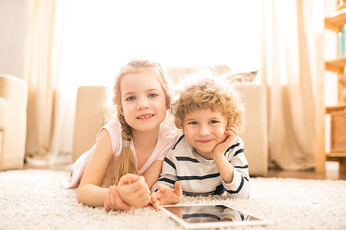 Cute happy girl and her adorable brother with tablet lying on the floor of living-room at leisure