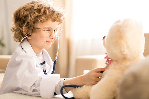 Cute little blond boy with curly hair examining soft white teddybear with stethoscope while playing at home or kindergarten