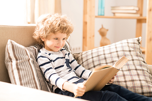Cute and clever young boy in casualwear sitting in comfortable armchair and reading book of tales