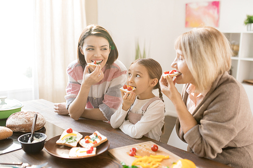 Happy women and little girl eating tasty self-made sandwiches with cheese and fresh vegetables for breakfast