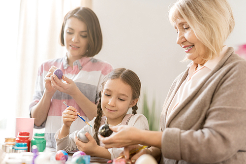 Mature woman showing her granddaughter one of Easter eggs while painting them before holiday