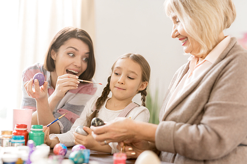 Playful young woman pointing at her daughter with paintbrush while having fun and painting eggs for Easter