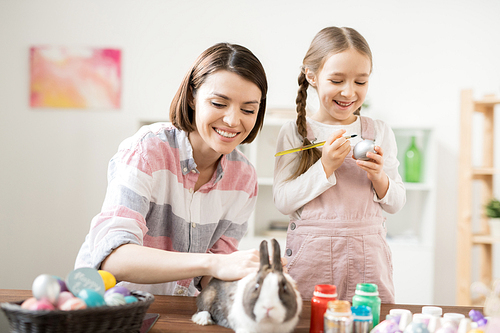 Happy young woman cuddling cute Easter bunny while her little daughter painting eggs for holiday