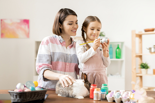 Young cheerful woman cuddling bunny and looking at her little daughter painting Easter eggs