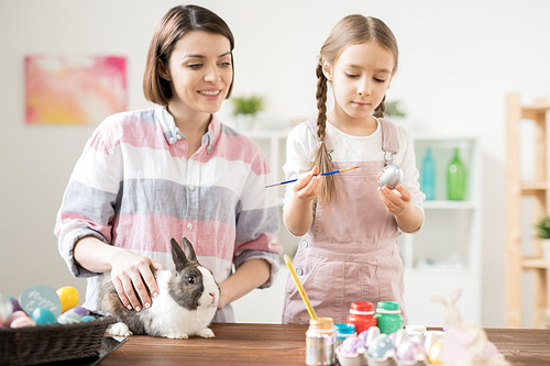 Adorable girl painting Easter egg in silver color with her mom standing near by and cuddling fluffy rabbit