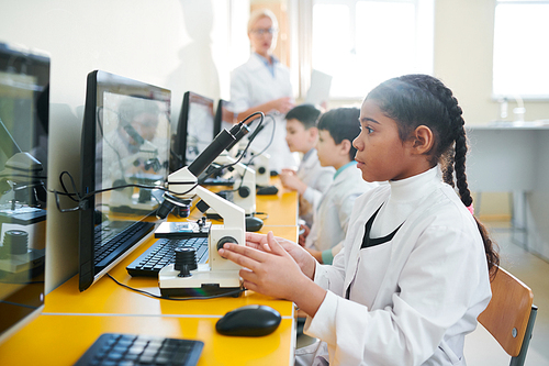 Row of little schoolkids in whitecoats sitting in classroom along desk and studying substances in microscope