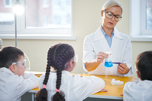Female teacher of chemistry in whitecoat and eyeglasses explaining her students chemical reaction of two substances
