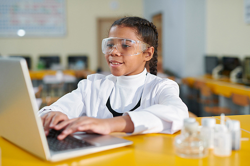 Youthful schoolgirl in protective eyeglasses and whitecoat sitting by desk in front of laptop at lesson