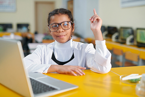 Cute secondary school pupil in whitecoat and eyeglasses raising her hand while sitting by desk at lesson