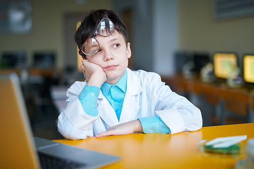 Funny schoolboy with his eyeglasses on forehead sitting by desk and thinking of new subject of lesson