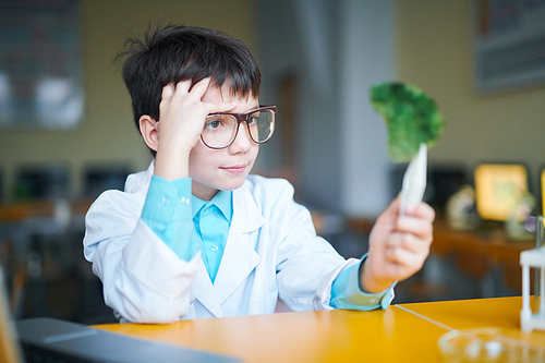 Pensive clever schoolboy in eyeglasses and whitecoat looking at green crystal while sitting by desk at lesson