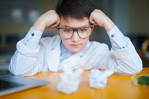 Pensive schoolboy in eyeglasses and whitecoat looking at white crystals while carrying out laboratory assignment