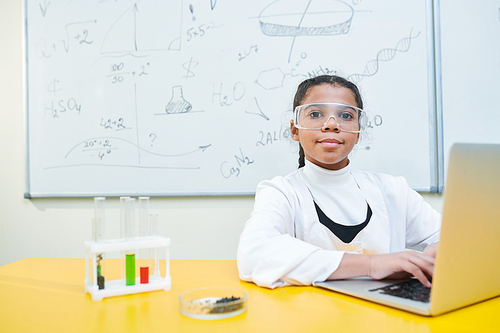 Clever and diligent schoolgirl in protective eyewear and whitecoat sitting by desk in front of laptop at lesson