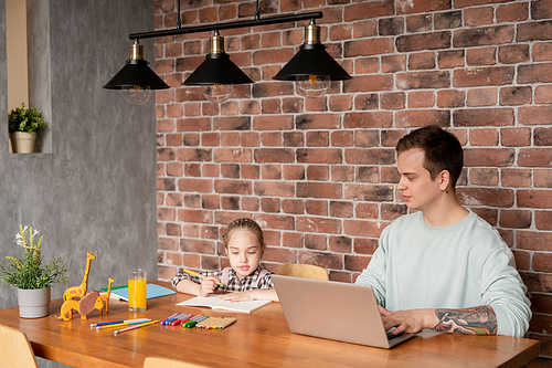 Content hipster young father with tattoo on arm sitting at wooden table and working with laptop at home while his daughter drawing picture with pencils