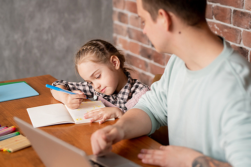Content cute little daughter with braid sitting at table and showing drawing in notebook to father working with laptop at home