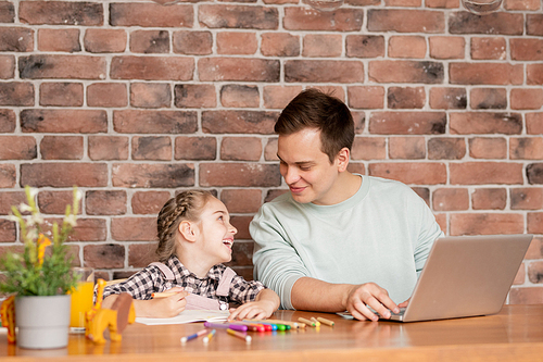 Cheerful excited young father and beautiful daughter with braid sitting at table in loft flat and working on school project together using multi-colored pencils and laptop