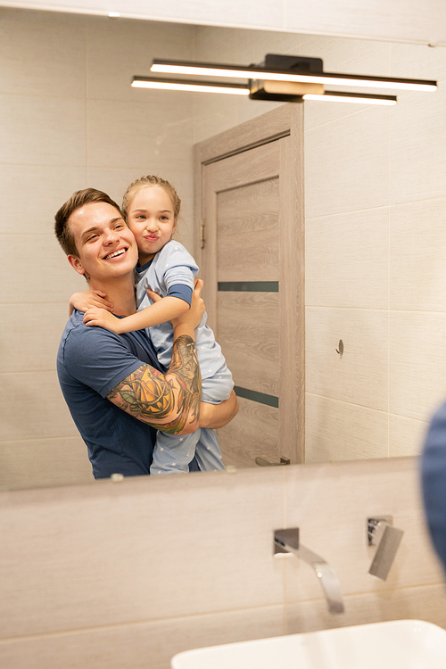 Happy excited young single father with tattoo on arm holding preschool daughter and looking into mirror together in bathroom