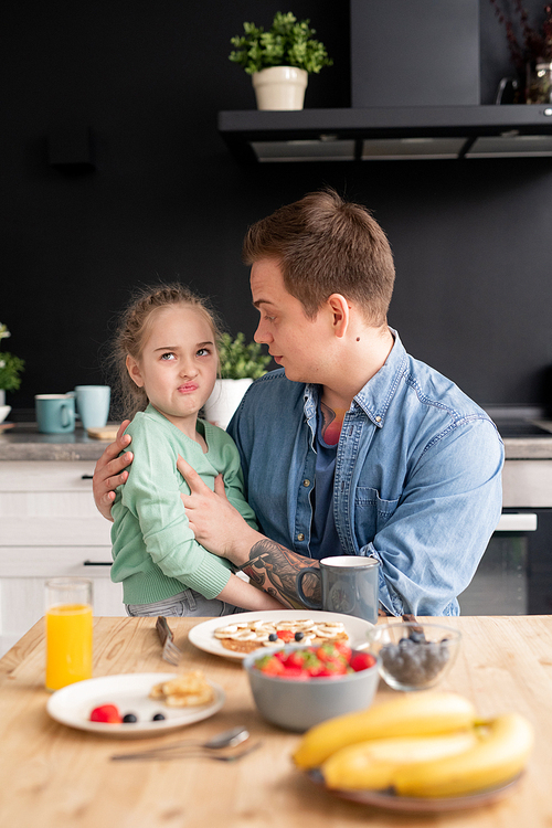 Picky daughter sitting on fathers knees and grimacing while refusing to eat breakfast made by father