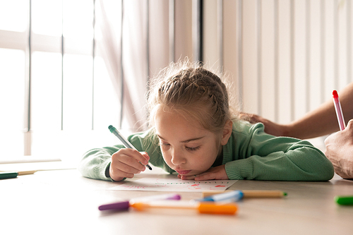 Serious  pretty girl with braided hair lying on floor and coloring picture with felt-tip pens while her father supporting her creativity and stroking her back