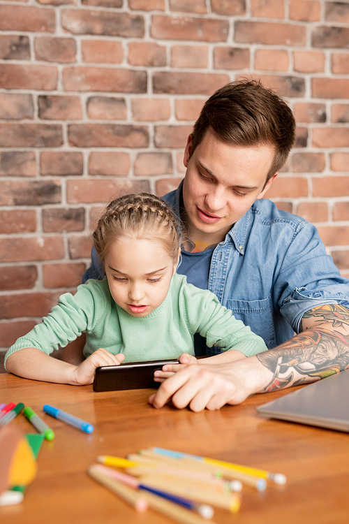 Content handsome young father and his curious daughter sitting at table and using mobile app on smartphone together