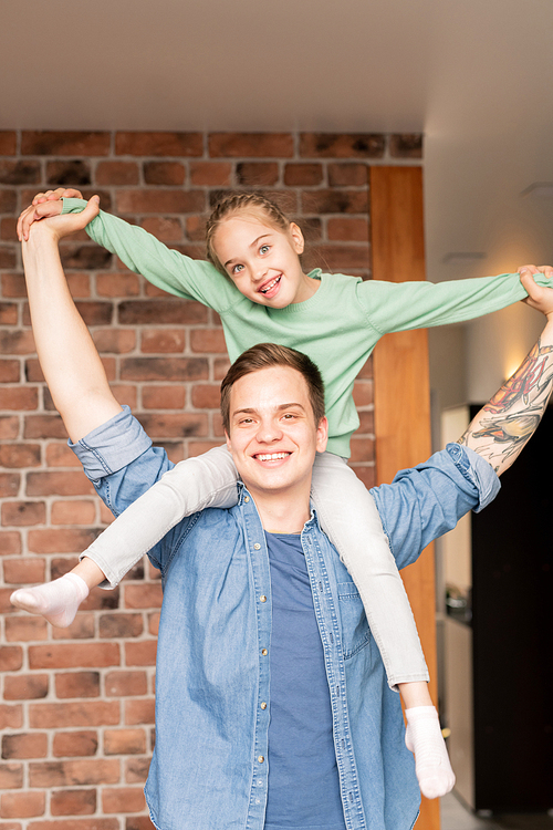 Positive playful young father with tattoo on arm and daughter with braided hair having fun together at home, excited girl sitting on neck of father