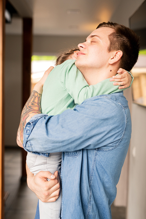Calm loving hipster young father with tattoo on arm keeping eyes closed while hugging adorable daughter and consoling her