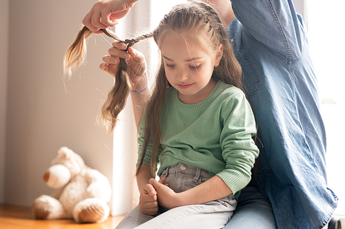 Careful young father in denim shirt braiding hair of daughter, upset girl sitting on fathers knees and looking down