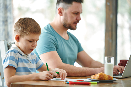 Cute little boy drawing with green highlighter by breakfast while his father networking on background