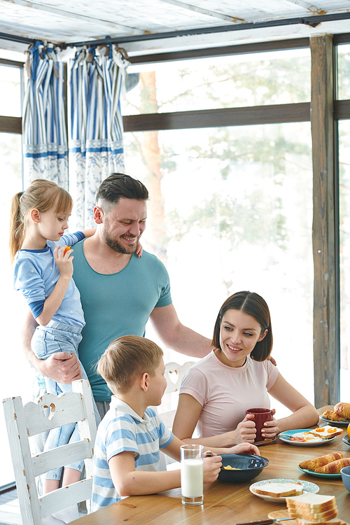 Happy mother and her little son having talk by table while young father with daughter standing near by