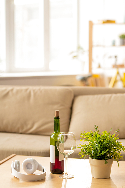 White headphones, empty wineglass, bottle of red wine and domestic plant in flowerpot on wooden table on background of couch
