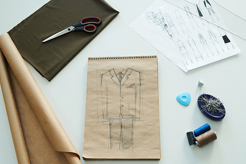 Overview of sketch of male suit in notepad, bunch of pins, thimble, colorful spools, piece of chalk, scissors, brown fabric and rolled paper on desk