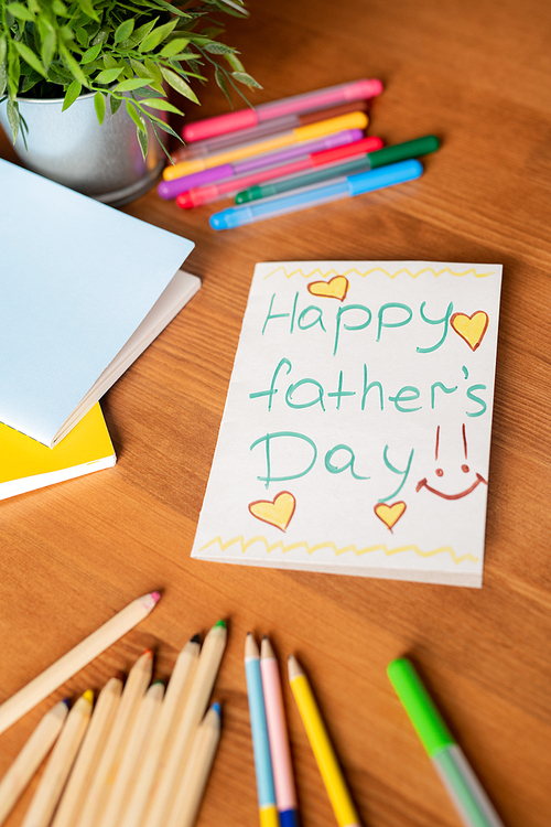 Close-up of handmade fathers day card with yellow hearts and colorful pencils and felt-tip pens on wooden table with potted plant and notebooks