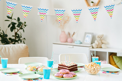 Table served for home birthday party for kids with cake with candles, drinks in blue paper glasses, popcorn, donuts and merengues