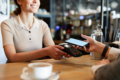 Young waitress standing by bar counter while holding electronic payment machine and waiting for client with smartphone paying for drink