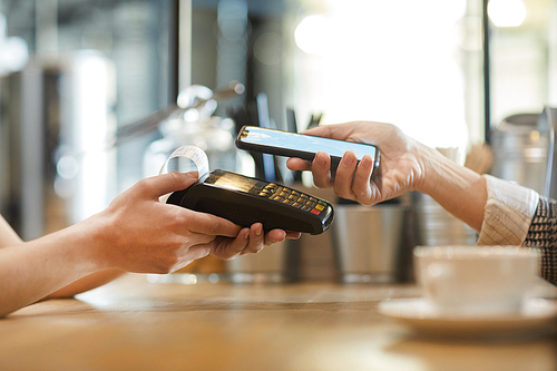Side view of hands of waitress with electronic device and those of client with smartphone during contactless payment in cafe