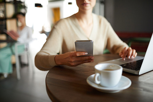 Young mobile contemporary businesswoman holding smartphone while texting or scrolling during network in front of laptop