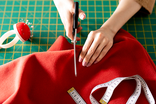 Hands of young seamstress with scissors cutting red textile while working over new coat for fashion collection