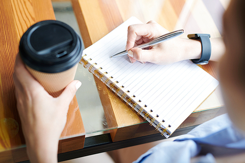 Close-up of unrecognizable woman sitting at table and drinking coffee while writing down plans and ideas in notepad