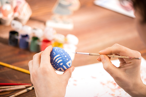 Human hand holding paintbrush while making white ornaments on blue egg while preparing for Easter