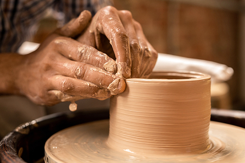 Hands of artisan holding rotating workpiece of clay pot while working by pottery wheel in his workshop