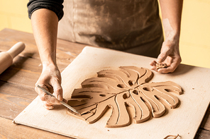 Creative craftswoman with pencil pointing at clay item in shape of leaf on wooden board during work over its design