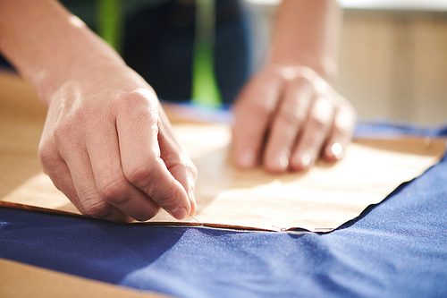Hand of young seamstress pinning up paper pattern on blue fabric before encircling it