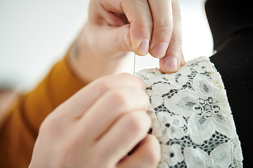 Hand of professional tailor putting pin through white lace while working over wedding dress in studio of fashion