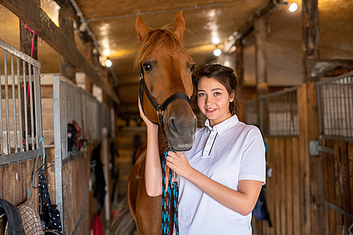 Pretty young smiling brunette keeping one hand by muzzle of purebred brown mare while standing in stable