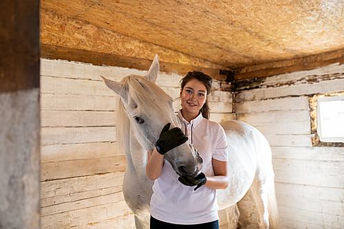 Happy young active woman in white shirt and black leather gloves embracing muzzle of purebred mare in stable