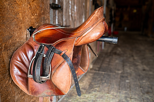Brown shabby leather saddle with black bridles hanging on steel bar stuck out of wall inside stable