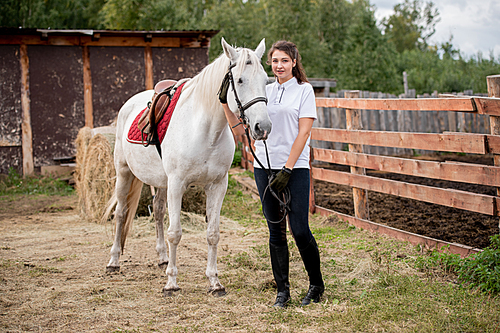 young active woman in jeans, polo shirt and boots looking at you while chilling out with white purebred racehorse in rural