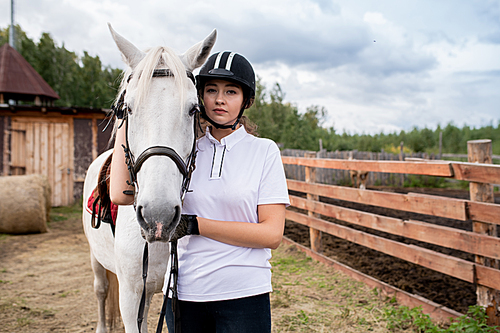 Young sportswoman in activewear and equestrian helmet standing by white purebred racehorse in front of camera