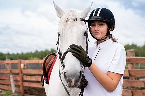 calm young woman in white polo shirt and equestrian outfit embracing purebred horse while training in rural